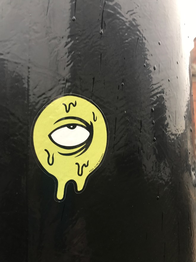 Green eyeball sticker on a pole. The eyeball is rolled up in the socket. Panic is the mood.